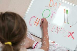 Top view of Little school girl with chickenpox drawing on white board in kids' room, antiseptic cream applied to face and body. Chalkboard and toys background. photo