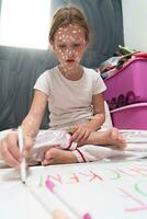 Little school girl with chickenpox drawing on white board in kids' room, antiseptic cream applied to face and body. Chalkboard and toys background. photo