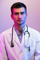 Coronavirus covid-19 danger alert Portrait of hero in white coat. Cheerful smiling young doctor with stethoscope in medical hospital standing against blue and pink background. photo