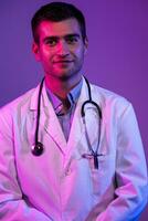 Coronavirus covid-19 danger alert Portrait of hero in white coat. Cheerful smiling young doctor with stethoscope in medical hospital standing against blue and pink background. photo