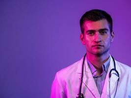 Coronavirus covid-19 danger alert Portrait of hero in white coat. Cheerful smiling young doctor with stethoscope in medical hospital standing against blue and pink background. photo