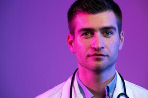 Coronavirus covid-19 danger alert Portrait of hero in white coat. Cheerful smiling young doctor with stethoscope in medical hospital standing against blue and pink background. photo