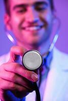 Portrait of hero in white coat. Cheerful smiling young doctor with stethoscope in medical hospital standing against blue and pink background. Coronavirus covid-19 danger alert photo