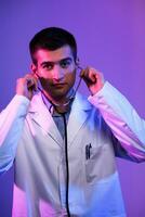 Portrait of a hero in a white coat. Cheerful smiling young doctor with stethoscope in medical hospital standing on blue and pink background. Warning about the danger of the coronavirus covid-19 photo