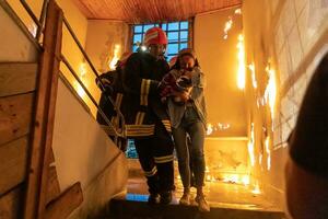 valiente bombero desciende escalera de un ardiente edificio y sostiene salvado niña en su brazos. abierto fuego y uno bombero en el antecedentes. foto