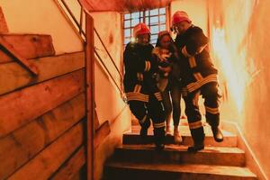 valiente bombero desciende escalera de un ardiente edificio y sostiene salvado niña en su brazos. abierto fuego y uno bombero en el antecedentes. foto