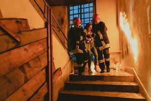 valiente bombero desciende escalera de un ardiente edificio y sostiene salvado niña en su brazos. abierto fuego y uno bombero en el antecedentes. foto