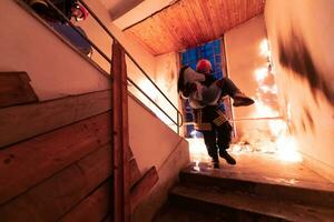 valiente bombero desciende escalera de un ardiente edificio y sostiene salvado niña en su brazos. abierto fuego y uno bombero en el antecedentes. foto