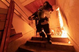 Brave Fireman Descends Stairs of a Burning Building and Holds Saved Girl in His Arms. Open fire and one Firefighter in the Background. photo