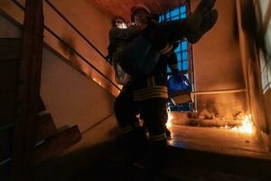 Brave Fireman Descends Stairs of a Burning Building and Holds Saved Girl in His Arms. Open fire and one Firefighter in the Background. photo