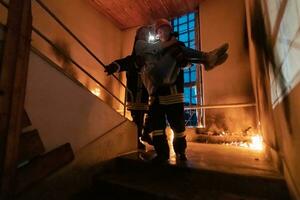 Brave Fireman Descends Stairs of a Burning Building and Holds Saved Girl in His Arms. Open fire and one Firefighter in the Background. photo