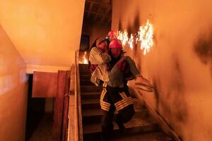 Brave Fireman Descends Stairs of a Burning Building and Holds Saved Girl in His Arms. Open fire and one Firefighter in the Background. photo