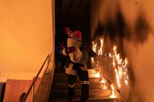 Brave Fireman Descends Stairs of a Burning Building and Holds Saved Girl in His Arms. Open fire and one Firefighter in the Background. photo