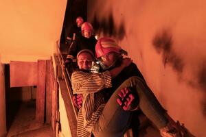 Brave Fireman Descends Stairs of a Burning Building and Holds Saved Girl in His Arms. Open fire and one Firefighter in the Background. photo