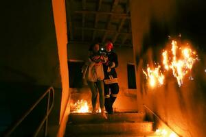 Brave Fireman Descends Stairs of a Burning Building and Holds Saved Girl in His Arms. Open fire and one Firefighter in the Background. photo