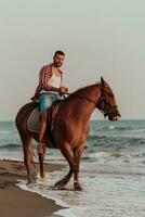 A modern man in summer clothes enjoys riding a horse on a beautiful sandy beach at sunset. Selective focus photo