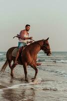 A modern man in summer clothes enjoys riding a horse on a beautiful sandy beach at sunset. Selective focus photo