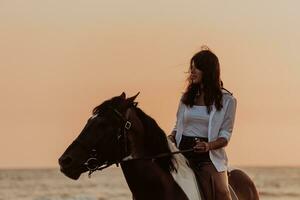 Woman in summer clothes enjoys riding a horse on a beautiful sandy beach at sunset. Selective focus photo