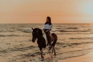 Woman in summer clothes enjoys riding a horse on a beautiful sandy beach at sunset. Selective focus photo