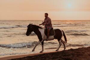 A modern man in summer clothes enjoys riding a horse on a beautiful sandy beach at sunset. Selective focus photo