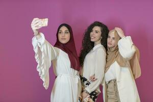 group of beautiful muslim women two of them in fashionable dress with hijab using mobile phone while taking selfie picture isolated on pink background representing modern islam fashion technology photo