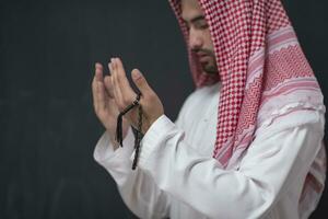 A young Arabian man in traditional clothes making a traditional prayer to God keeps his hands in praying gesture in front of a black background photo