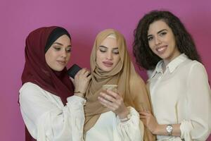 Group of beautiful muslim women two of them in fashionable dress with hijab using mobile phone isolated on pink background representing modern islam fashion technology and ramadan kareem concept photo