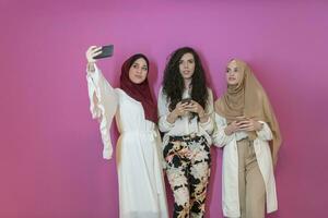 grupo de hermosa musulmán mujer dos de ellos en de moda vestir con hijab utilizando móvil teléfono mientras tomando selfie imagen aislado en rosado antecedentes representando moderno islam Moda tecnología foto