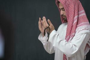 A young Arabian man in traditional clothes making a traditional prayer to God keeps his hands in praying gesture in front of a black background photo