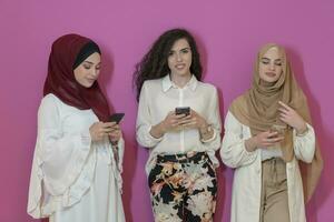 Group of beautiful muslim women two of them in fashionable dress with hijab using mobile phone isolated on pink background representing modern islam fashion technology and ramadan kareem concept photo