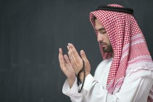 A young Arabian man in traditional clothes making a traditional prayer to God keeps his hands in praying gesture in front of a black background photo