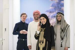 Group portrait of young Muslim people women in fashionable hijab dress with three Arabian men using smartphones at modern bright home photo