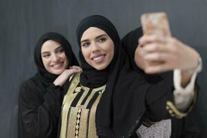 grupo de joven hermosa musulmán mujer en de moda vestir con hijab utilizando teléfono inteligente mientras tomando selfie imagen en frente de negro antecedentes foto