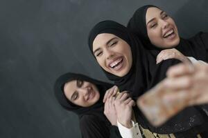 grupo de joven hermosa musulmán mujer en de moda vestir con hijab utilizando teléfono inteligente mientras tomando selfie imagen en frente de negro antecedentes foto