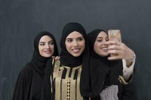 Group of young beautiful muslim women in fashionable dress with hijab using smartphone while taking selfie picture in front of black background photo