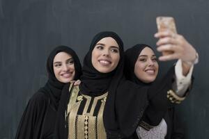 grupo de joven hermosa musulmán mujer en de moda vestir con hijab utilizando teléfono inteligente mientras tomando selfie imagen en frente de negro antecedentes foto