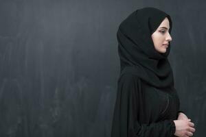 Portrait of beautiful muslim woman in fashionable dress with hijab in front of black chalkboard representing modern islam fashion and ramadan kareem concept photo