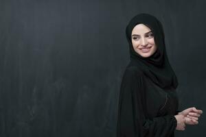 Portrait of beautiful muslim woman in fashionable dress with hijab in front of black chalkboard representing modern islam fashion and ramadan kareem concept photo