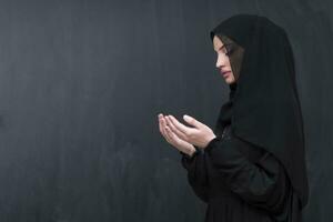 Portrait of beautiful muslim woman in fashionable dress with hijab making traditional prayer to God keeps hands in praying gesture isolated on black background photo
