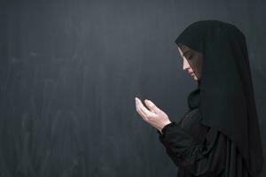 Portrait of beautiful muslim woman in fashionable dress with hijab making traditional prayer to God keeps hands in praying gesture isolated on black background photo