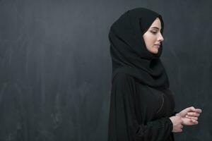 Portrait of beautiful muslim woman in fashionable dress with hijab in front of black chalkboard representing modern islam fashion and ramadan kareem concept photo