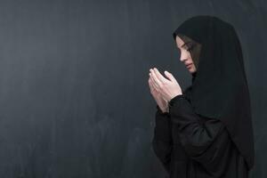 Portrait of beautiful muslim woman in fashionable dress with hijab making traditional prayer to God keeps hands in praying gesture isolated on black background photo