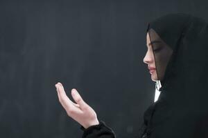 Portrait of beautiful muslim woman in fashionable dress with hijab making traditional prayer to God keeps hands in praying gesture isolated on black background photo