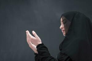 Portrait of beautiful muslim woman in fashionable dress with hijab making traditional prayer to God keeps hands in praying gesture isolated on black background photo