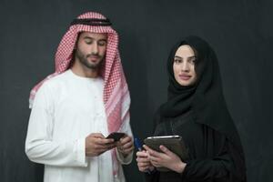 Young muslim business couple in fashionable hijab dress using smartphone and tablet in front of black background photo