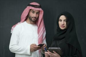 Young muslim business couple in fashionable hijab dress using smartphone and tablet in front of black background photo