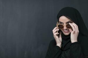 Young modern muslim businesswoman using smartphone wearing sunglasses and hijab clothes in front of black chalkboard photo