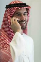 Young modern Muslim Arabian businessmen wearing traditional clothes while using smartphones at home. High-quality photo