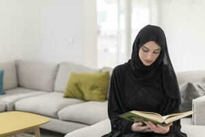 joven tradicional musulmán mujer leyendo Corán en el sofá antes de iftar cena durante un Ramadán banquete a hogar foto