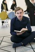 Young muslim man sitting on the floor while reading holy book Quran before iftar dinner during a ramadan feast at home photo